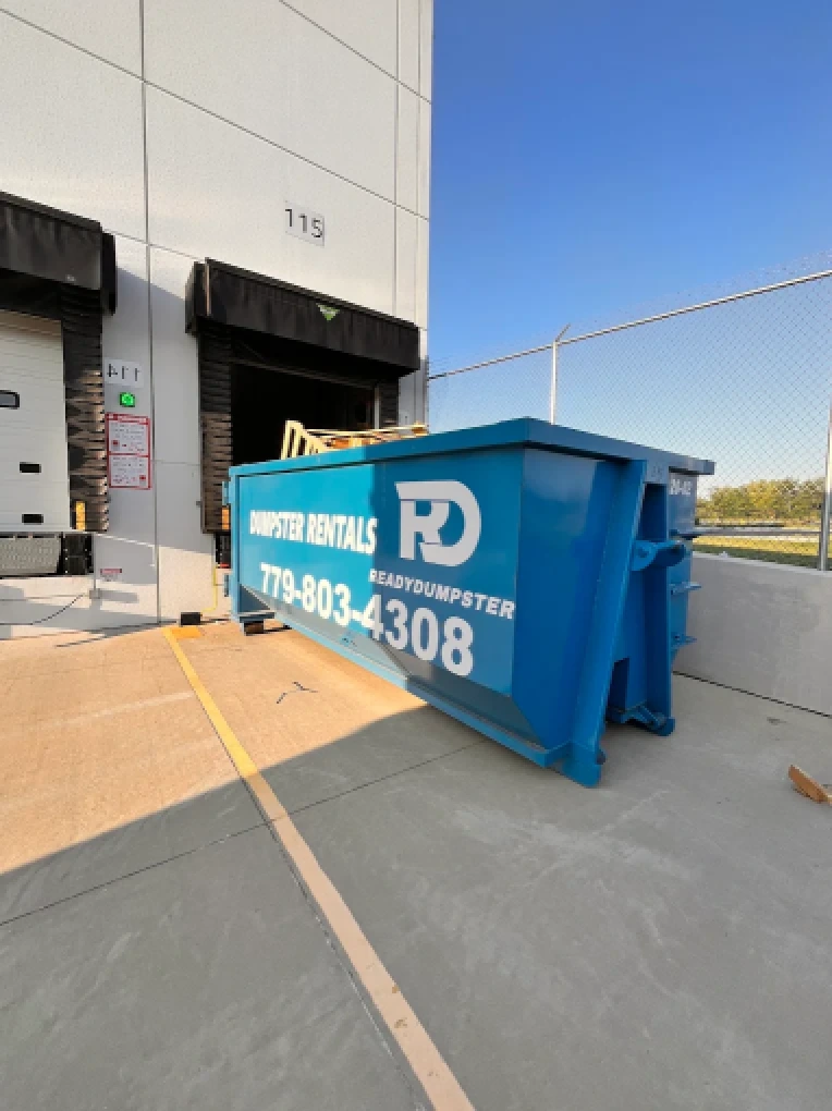 Large blue commercial dumpster staged outdoors on concrete near a commercial loading dock structure.