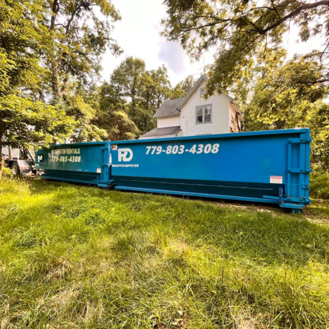 Blue roll-off dumpster parked on a grassy yard next to a two-story residential house.