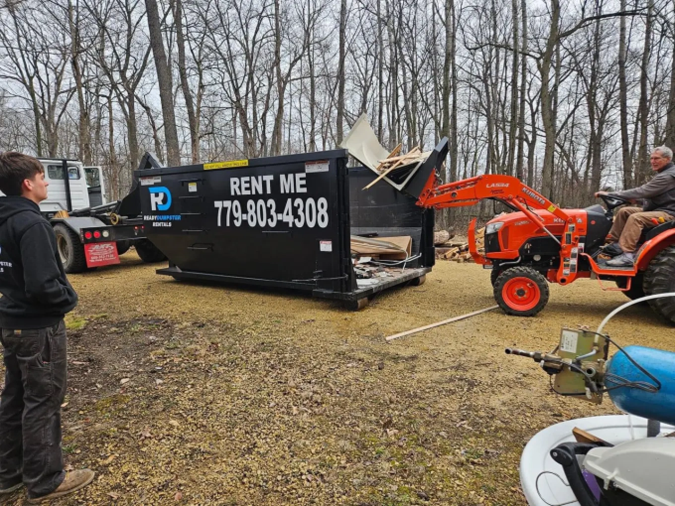 Black roll-off dumpster on a gravel construction site being loaded with wood debris using an orange tractor.